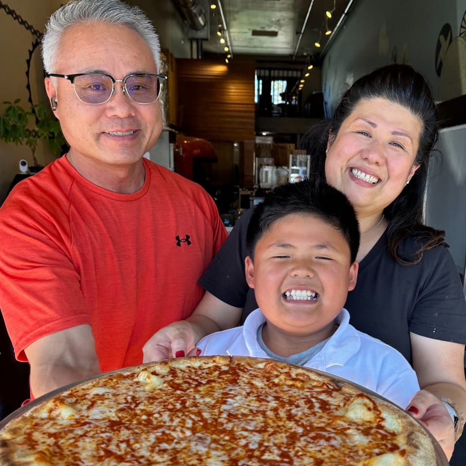 Three smiling people, two adults and a child, holding a large cheese pizza from Pj's Pizza Co.