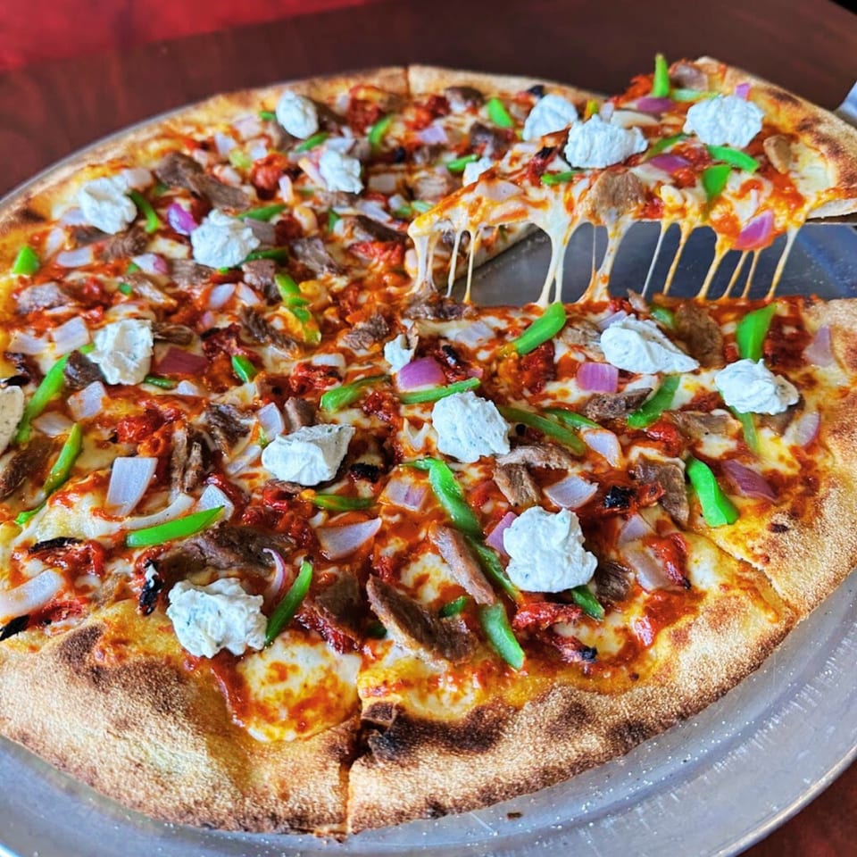 A slice of Pj's Pizza Co. pizza with steak, green peppers, onions, and ricotta is lifted, stretching cheese.