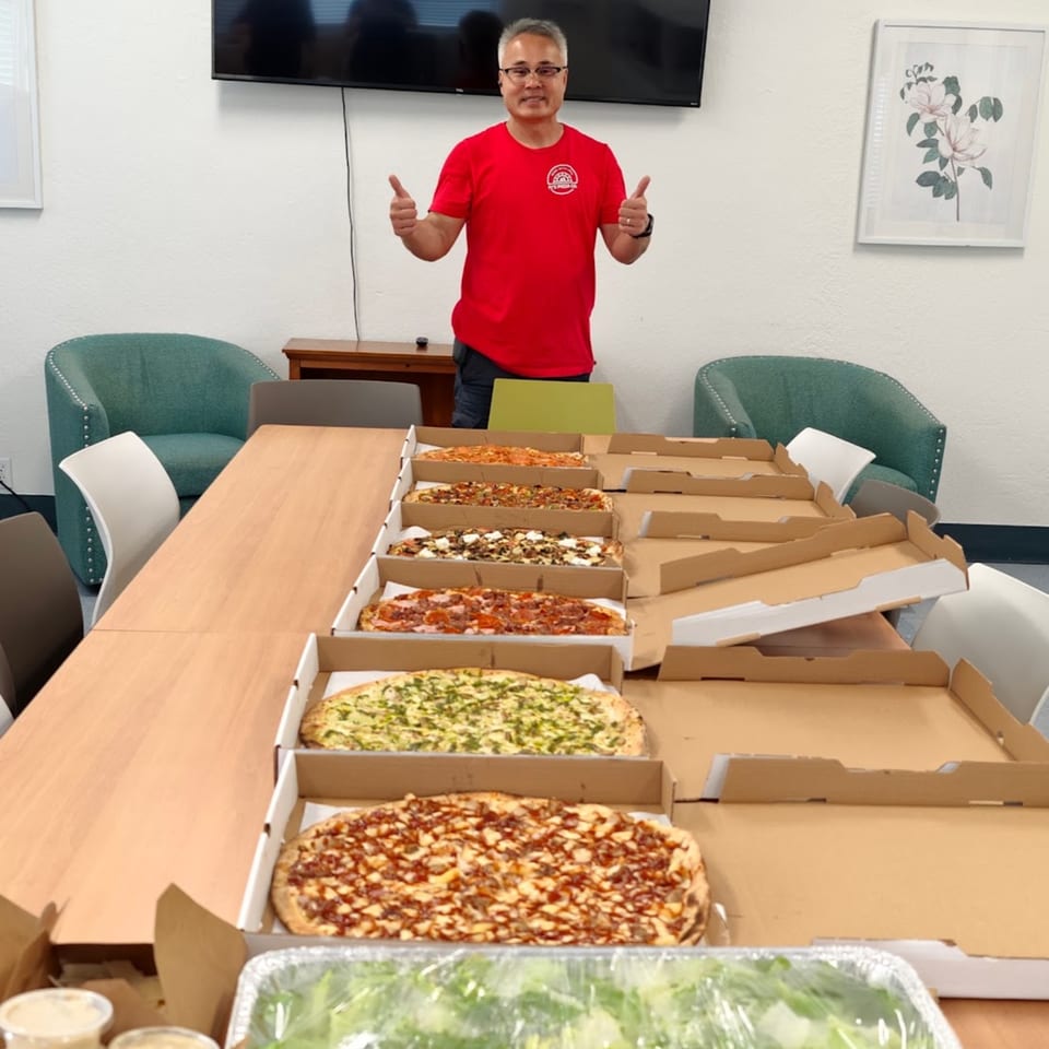 A man in a red Pj's Pizza Co. shirt gives thumbs up behind a table of pizzas.