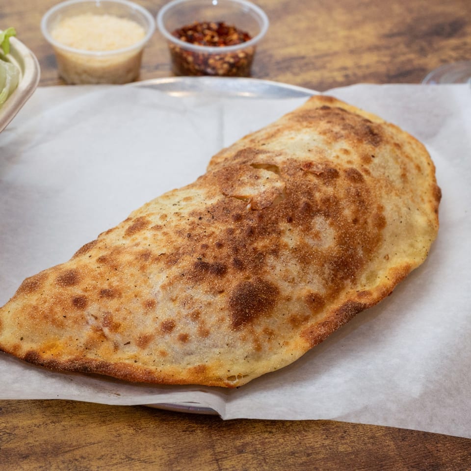 A golden-brown calzone from Pj's Pizza Co. on parchment paper, with parmesan and chili flakes in the background.