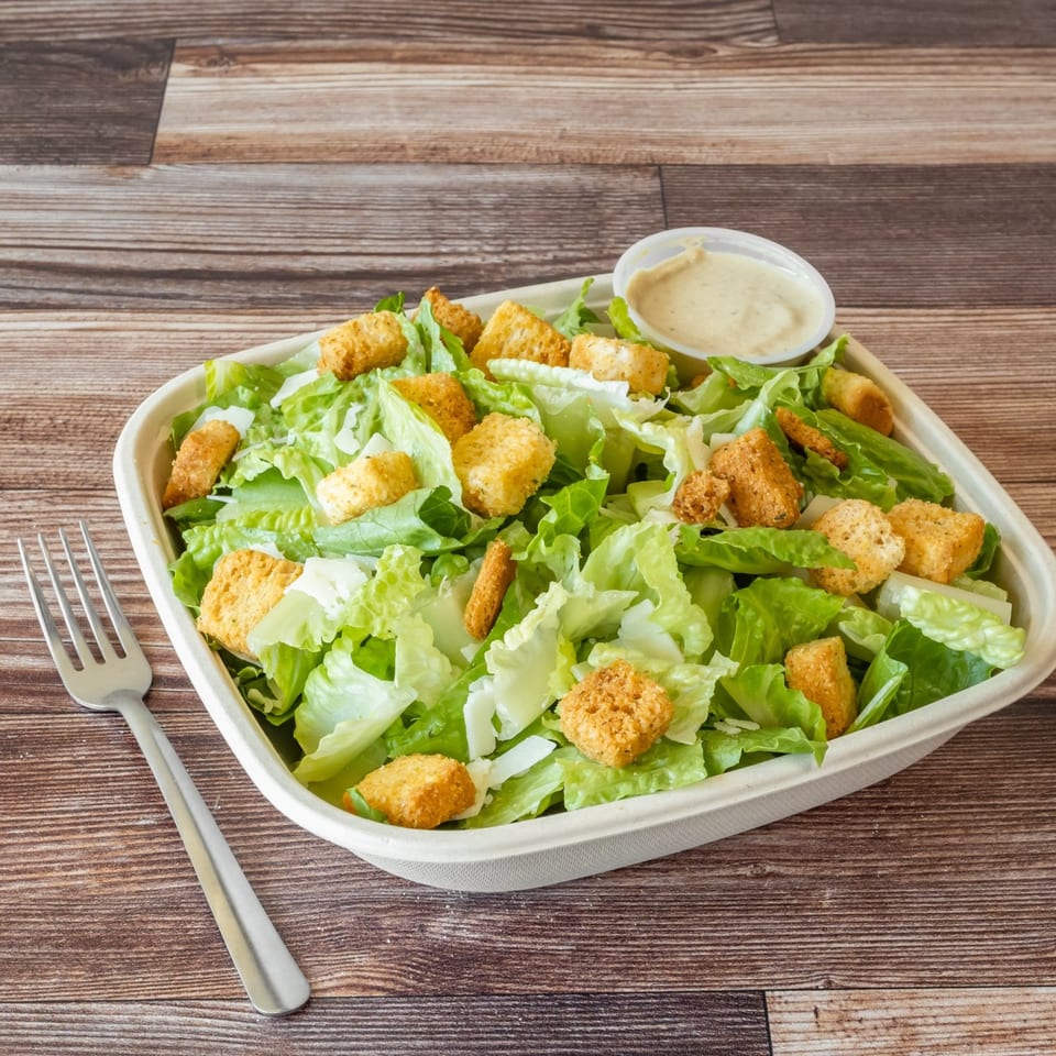 A fresh Caesar salad with croutons and dressing in a white container, next to a fork.